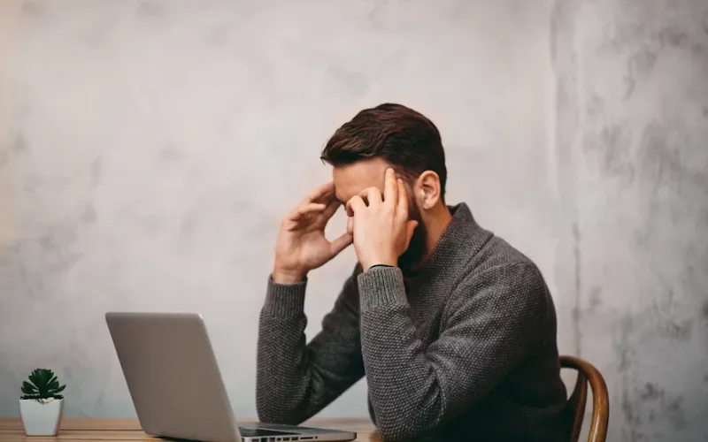 Man sitting at a desk holding his head while looking at a laptop, showing stress, anxiety, and mental overload.