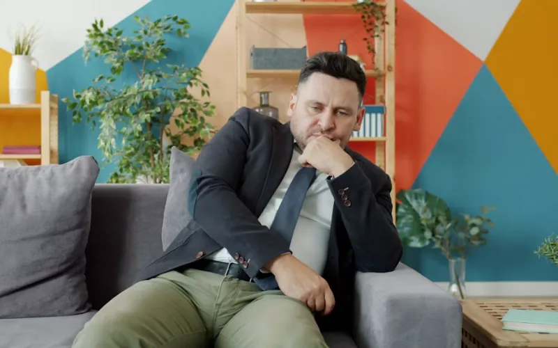 Thoughtful man sitting on a sofa with his hand on his chin, appearing overwhelmed and deep in thought.